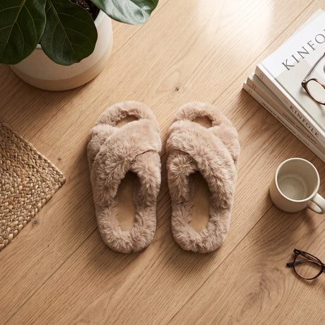 Beige fluffy crossover kids slippers laid flat on a wooden floor beside a mug and books
