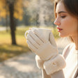 A woman in a cream knit sweater and beige fur trim gloves holds a steaming mug in an outdoor park setting