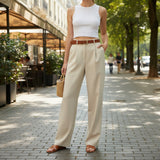 Woman in white top and beige pants standing on a sidewalk with trees and outdoor seating in the background