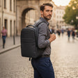 A man wearing a grey long-sleeve shirt and navy trousers walks on a cobblestone street while wearing a black backpack