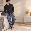 Man in a black button front pyjama top and blue check trousers leaning on a bed in a bright bedroom