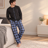 Man in a black button front pyjama top and blue check trousers leaning on a bed in a bright bedroom