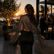 A woman stands with her back to the camera in a modern rooftop lounge, wearing a sheer black mesh shawl with long fringed sleeves over a black dress