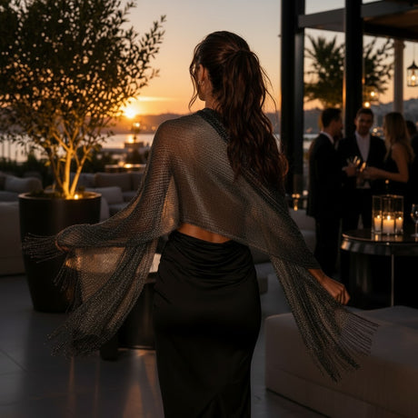 A woman stands with her back to the camera in a modern rooftop lounge, wearing a sheer black mesh shawl with long fringed sleeves over a black dress