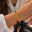 Wrist wearing a black cord bracelet with a flowing gold Arabic calligraphy charm near a black bag