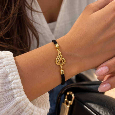 Wrist wearing a black cord bracelet with a flowing gold Arabic calligraphy charm near a black bag