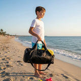 Young boy holding a black duffel bag on a sandy beach with ocean and clear sky in the background.