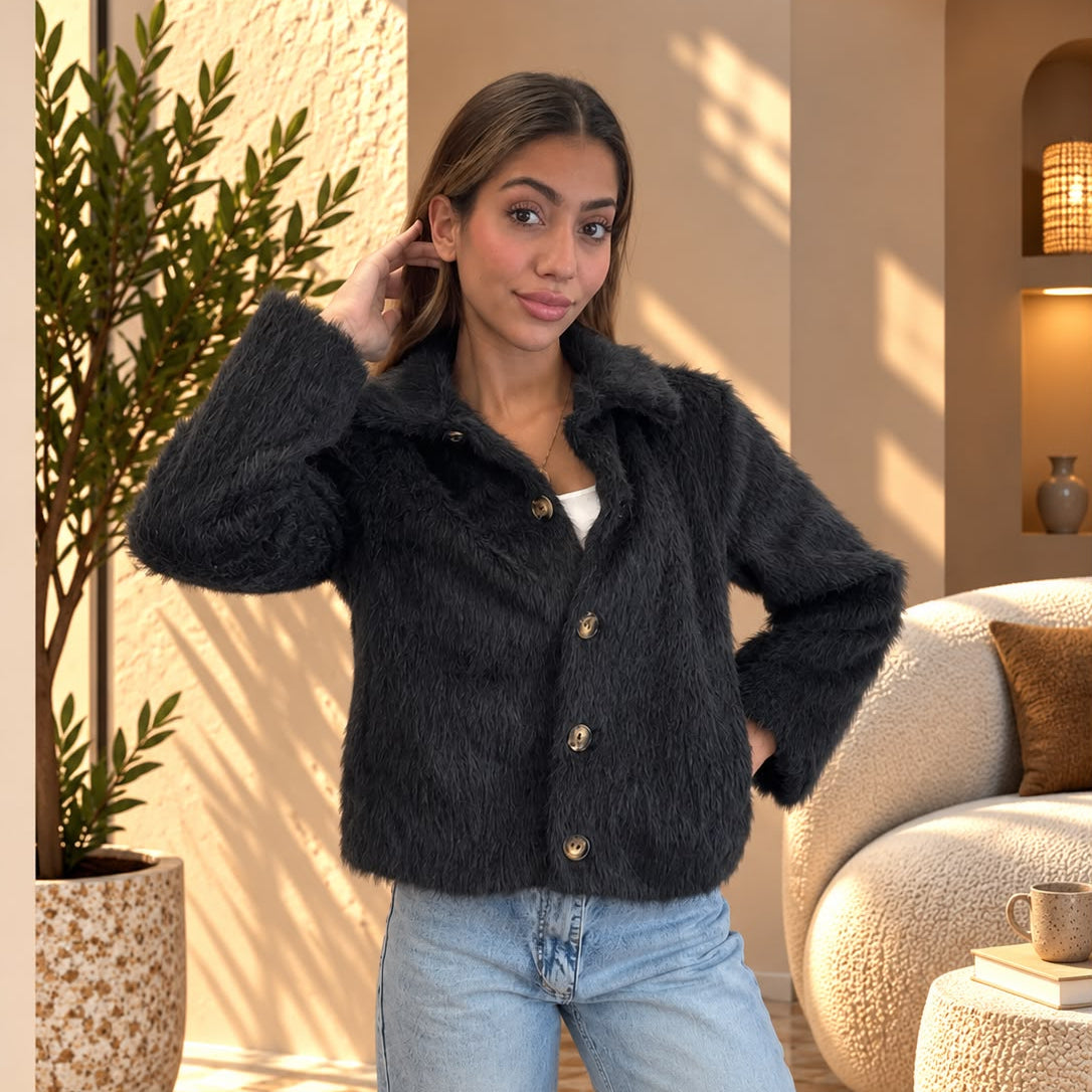 Fluffy black jacket buttoned up, one hand in hair, with light wash jeans