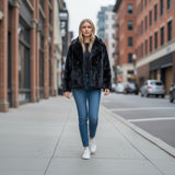 Woman walking on a city street wearing a black fur coat, blue jeans, and white sneakers