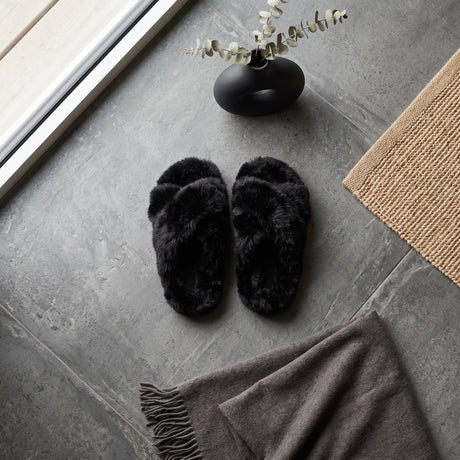 Black fluffy crossover kids slippers laid flat on a grey tiled floor by a window