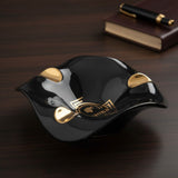 Wavy glossy black ashtray whose three rests shine in gold, set on a dark wood desk