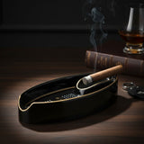 Lit cigar resting in a slim black oval ashtray beside a whiskey glass and books