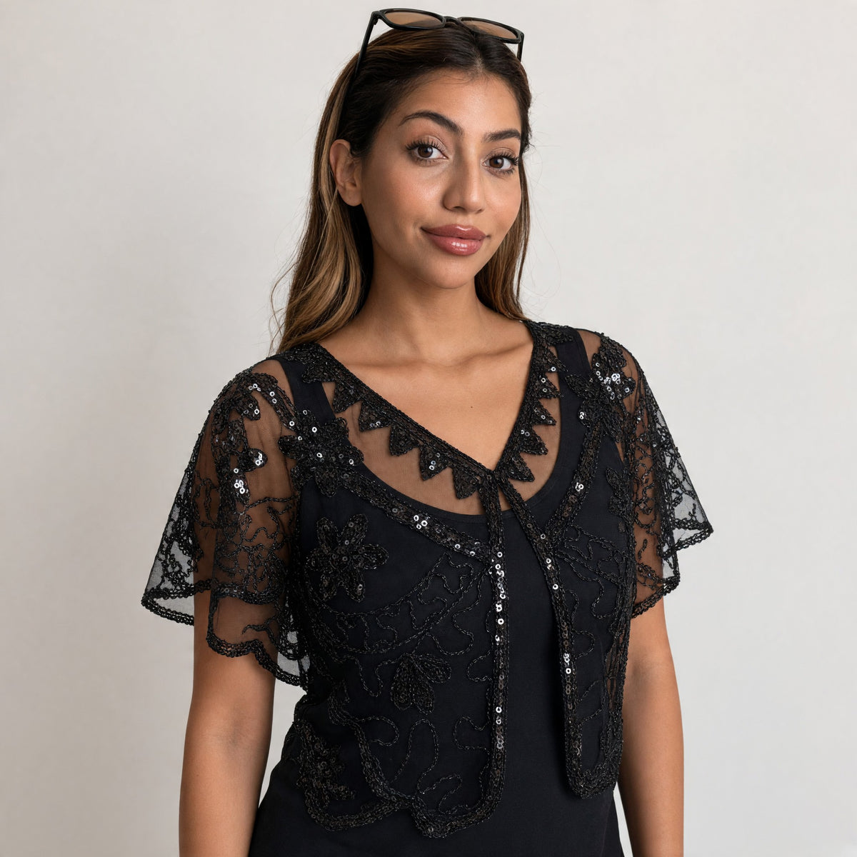 Woman wearing a black sheer mesh cape shrug with sequin and beaded floral embroidery over a black dress
