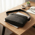 Black crossbody bag on a wooden desk with a laptop, glasses, and a cup of coffee