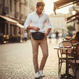 Man in a white shirt and beige pants wearing a black sling pack standing on a street