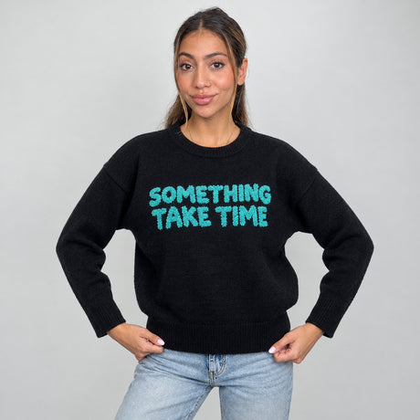 Black knit sweater with fuzzy teal slogan lettering across the chest, hands on hips