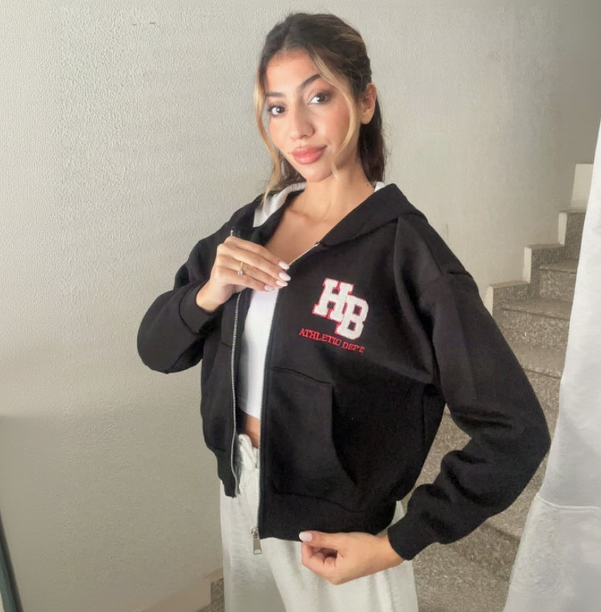 Black varsity patch zip hoodie worn open over a white top with grey joggers