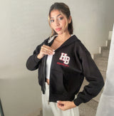 Black varsity patch zip hoodie worn open over a white top with grey joggers