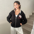 Woman in a black cropped zip hoodie with a varsity letter patch and grey joggers on a staircase