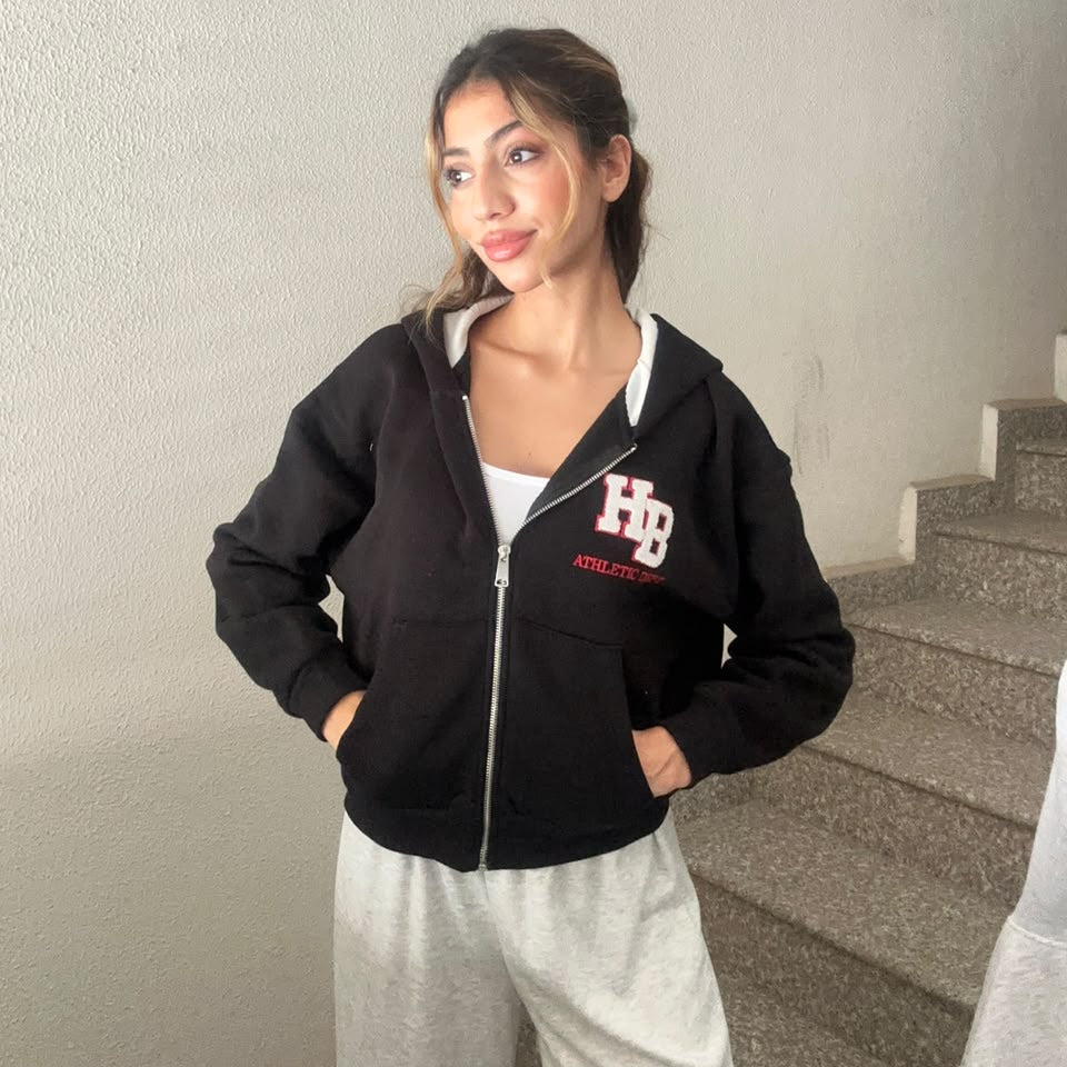Woman in a black cropped zip hoodie with a varsity letter patch and grey joggers on a staircase