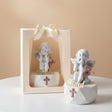 Matte white cherub with a rose pink cross on its base, beside a ribboned gift bag