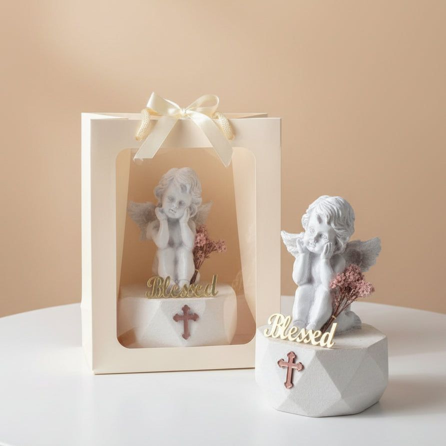 Matte white cherub with a rose pink cross on its base, beside a ribboned gift bag