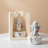 Matte white cherub with a rose pink cross on its base, beside a ribboned gift bag