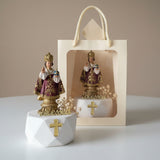 Crowned Infant of Prague in burgundy and gold beside a window gift bag