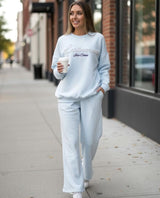 Woman in a light grey sweatshirt and sweatpants walking on a sidewalk, holding a coffee cup