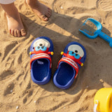 Top view of blue clogs on sand next to a spade and bare toddler feet