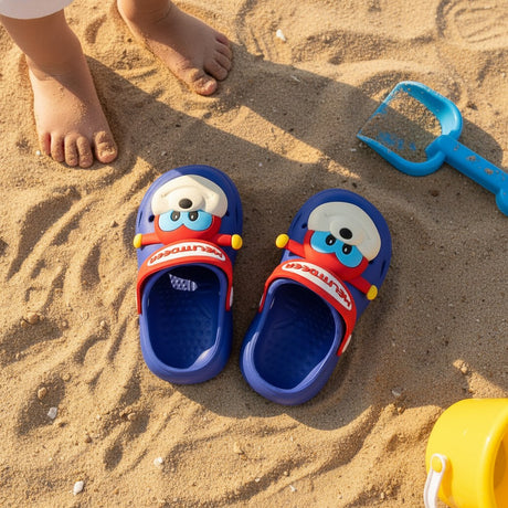 Top view of blue clogs on sand next to a spade and bare toddler feet