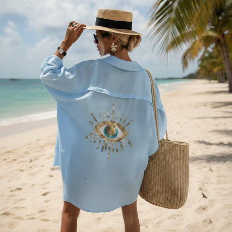 Woman on a beach wearing a light blue cover-up with a sun design, holding a straw bag.