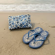 Blue floral pouch and flip-flops on a sandy beach with ocean waves in the background