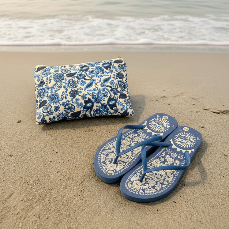 Blue floral pouch and flip-flops on a sandy beach with ocean waves in the background