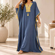 Woman in a blue batwing maxi kaftan with gold embroidered neckline and tassels, holding a clutch
