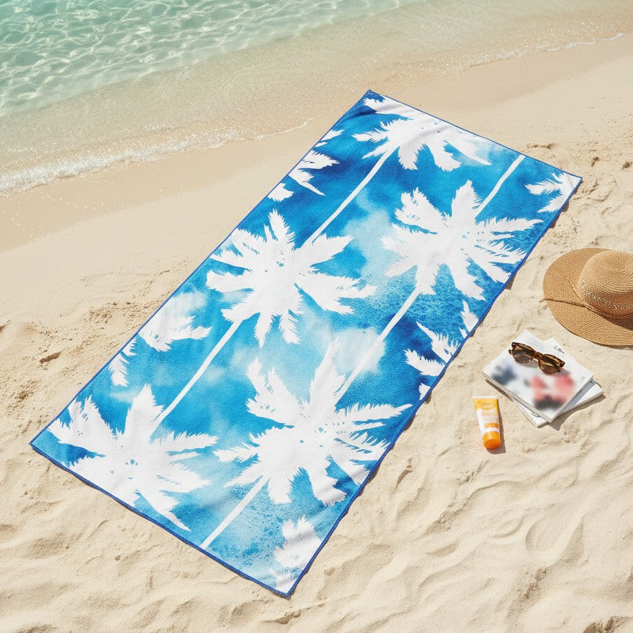A blue and white palm tree patterned beach towel is laid out on a sandy beach next to the ocean water, with a straw hat, sunglasses, and a small sunscreen bottle nearby.