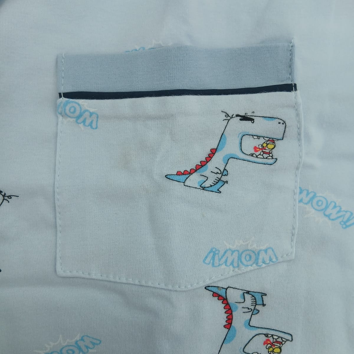 Chest pocket close up on a light blue kids pyjama top with cartoon dinosaur print and navy piping