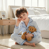Boy sitting on a bedroom rug holding a teddy bear in a light blue button front pyjama set