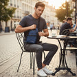 Man sitting at an outdoor cafe table with a blue sling bag, surrounded by urban scenery