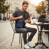 Man sitting at an outdoor cafe table with a blue sling bag, surrounded by urban scenery