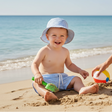 A baby wearing a light blue and white striped sun hat and matching swim trunks sits on a sandy beach playing with a green shovel