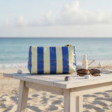 Blue and white striped beach bag on a wooden table with sunglasses and a bottle of sunscreen, set against a beach backdrop.