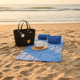 Black beach bag with a duck design, blue towels, and a hat on sand by the water