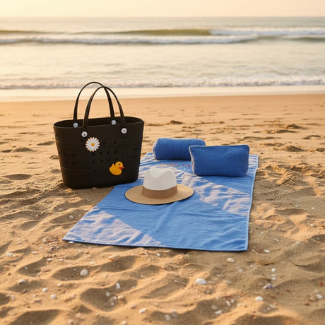 Black beach bag with a duck design, blue towels, and a hat on sand by the water