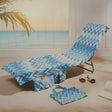 A beach-themed chaise lounge chair with a blue and white chevron pattern, set on a sandy beach with a matching tote bag, white slide sandals, and a palm leaf in the foreground.
