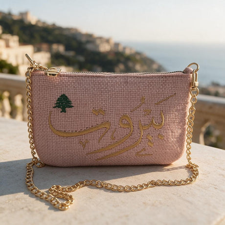 Pink textured bag resting on a white balustrade, Beirut stitched in gold script with a little cedar, coastline behind