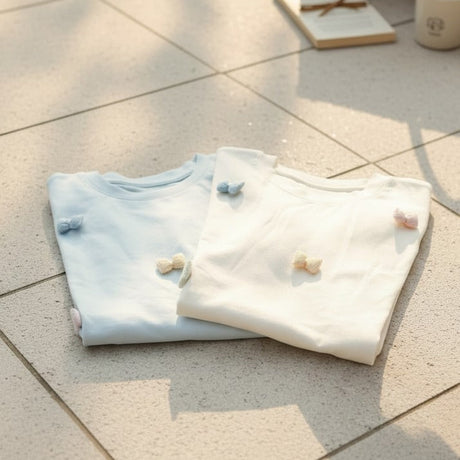 Light blue and cream bow detail tees folded side by side in the sun