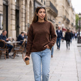 Woman in a brown brushed wool crew neck sweater and light jeans holding a coffee cup on a cafe lined street