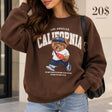 Woman in a brown oversized sweatshirt with a California print and a fuzzy walking teddy bear in a cap