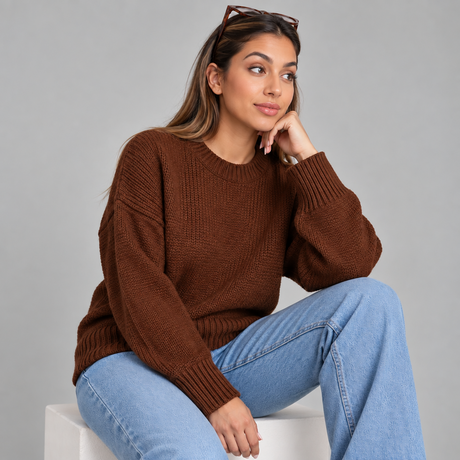 Woman in a brown chunky wool knit sweater and wide leg jeans seated on a white block, chin resting on her hand